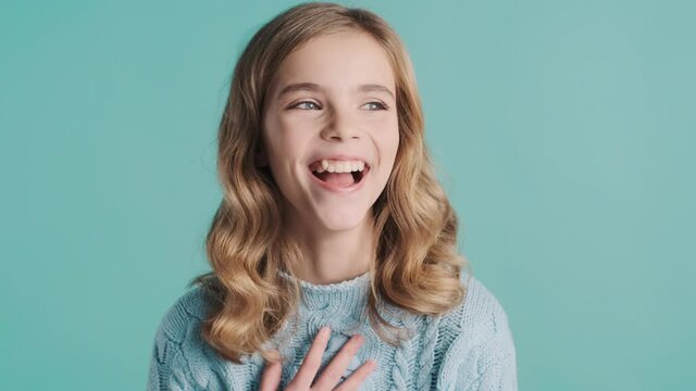 Pretty blond haired teenage girl dressed in cozy sweater looking happy smiling on camera over blue background