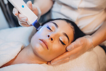 Obraz na płótnie Canvas Facial treatment procedure done to caucasian brunette with closed eyes at spa salon with apparatus