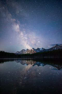 The Milky Way reflecting off of the calm waters of Herbert Lake while the town of Lake Louise illuminated the Canadian Rockies.