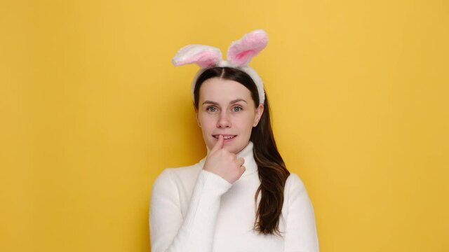Portrait of dreamful charming young woman in pink bunny fluffy ears, happy looking to camera, wears white jumper, standing isolated over yellow studio background. People emotion and easter concept