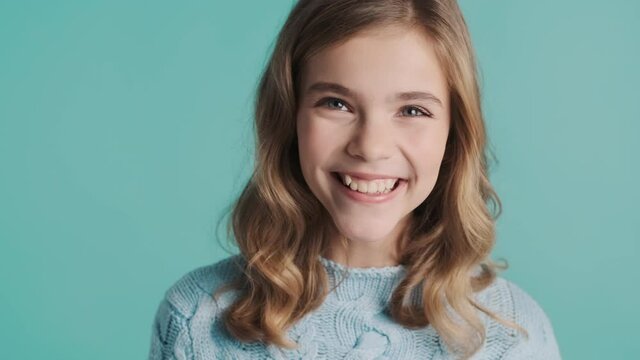 Cheerful blond teenage girl with wavy hair looking happy agreeing on camera over blue background. Pretty girl waving head yes