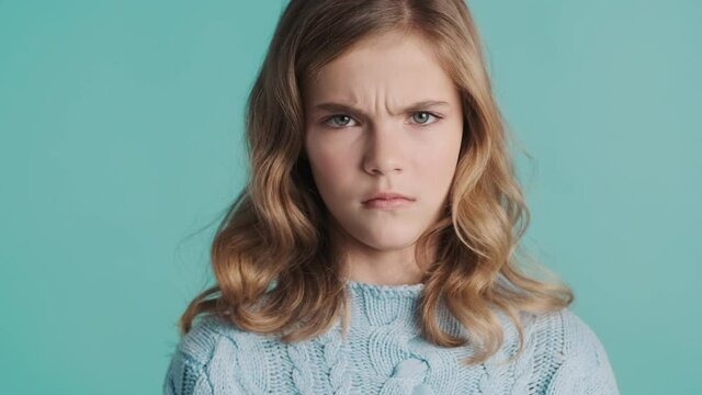 Serious blond teenager girl looking angry waving no on camera over blue background. Disagree expression