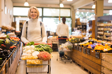Obraz premium Portrait of modern middle aged woman carrying purchases in trolley cart during shopping in grocery store