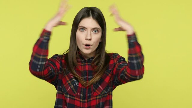 Bored exhausted brunette woman in checkered shirt touching her head and showing explosion, looking worried and shocked, deadline, professional burnout. Indoor studio shot isolated on yellow background