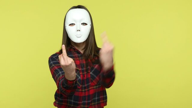 Rude aggressive unknown woman in checkered shirt threatening you hiding her face behind white mask, showing middle finger fuck you gesture, bullying. Indoor studio shot isolated on yellow background