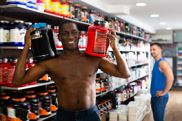 Glad athletic shirtless African man with jars of sports nutritional supplements showing muscular body in shop interior
