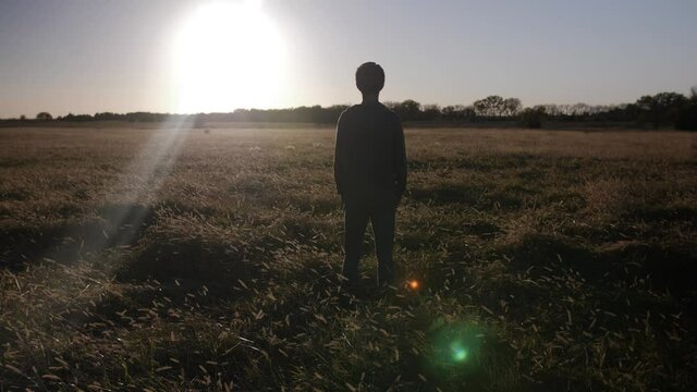 A young teenage boy stands in a Kansas field watching the sun set in slow motion.