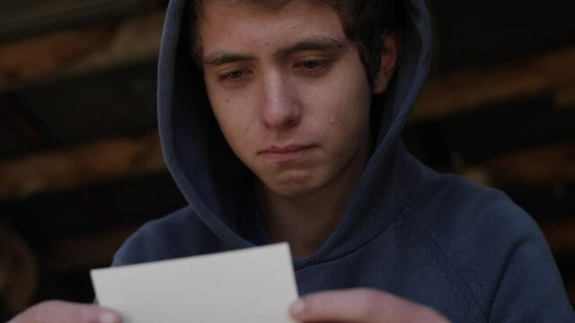 A young teenager sits in a garage looking at a picture of his dad. The teen boy smiles as a tear runs down his cheek.