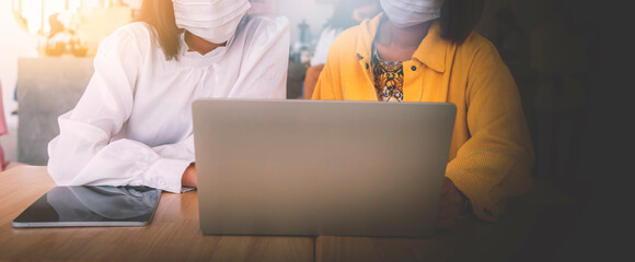 Two businesswoman wearing a mask looking on laptop and talk about business in cafe, cover banner design.