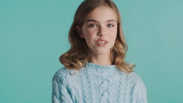 Charming blond teenage girl winking on camera sending air kiss and playing with eyebrow isolated on blue background. Flirty expression