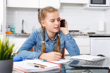 Confident teenage girl doing her home task using computer and her mobile phone in home interior