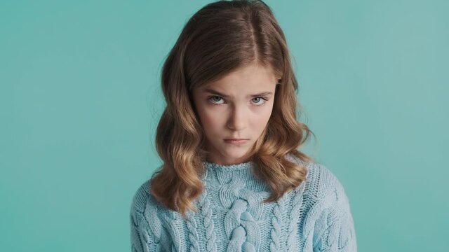 Upset blond teenager girl sadly looking from under the eyebrows in camera isolated on blue background. Guilty expression