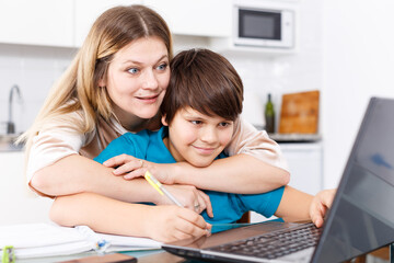 Portrait of smiling mother helping son with homework and having good time in kitchen
