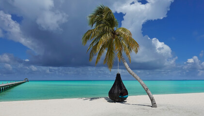 A palm tree bent over the sandy beach. A wicker swing is suspended from the trunk. Green leaves on a background of blue sky, clouds. There is a wooden walking path over the aquamarine ocean. Maldives