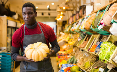 Obraz premium Focused African man working at farmer market, putting fresh organic pumpkin on showcase..