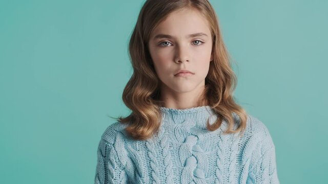 Upset blond teenage girl dressed in sweater feeling sad offendedly looking in camera over blue background