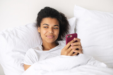 Happy young mixed-race woman with curly hair covered in a cozy soft blanket staying at home in the warm bed, texting chatting with friends on mobile phone, flirting, writing post for social media