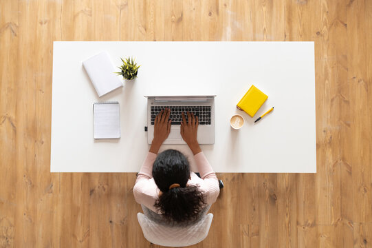 Overhead view of mixed-race ethnic woman or female freelancer with curly hair sitting at the white table desk with well-organized space, typing on the laptop, taking notes, modern home office concept