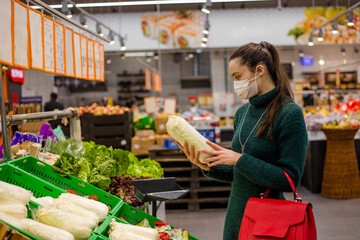 A woman wearing medical mask in a supermarket chooses fresh cabbage, a concept of agriculture and a...