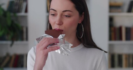 Young Caucasian woman eating chocolate bar close up indoors apartment background. Sensual brunette girl biting sweet candy with satisfaction on face. Unhealthy nutrition sugar addiction. Tasty food 