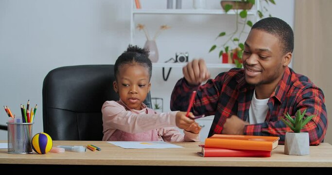 Happy family young father parent drawing coloring picture with pencils helping cute child daughter enjoying talk play sitting at table at home, dad and kid girl having fun learning in living room