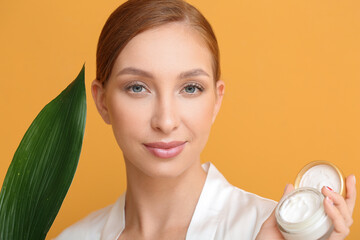 Beautiful young woman with facial cream and tropical leaf on color background, closeup