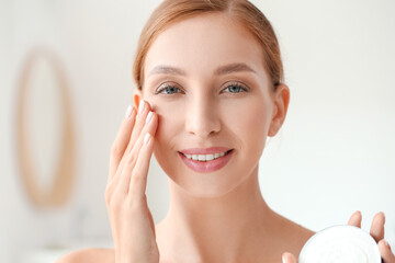 Beautiful young woman applying facial cream in bathroom, closeup