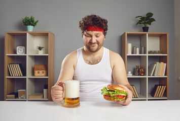 Funny athlete enjoying unhealthy food and alcohol. Happy guy eating big cheeseburger and drinking beer. Young man in sweatband and tank top sitting at table looking at glass mug and delicious burger