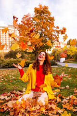 A girl tosses autumn leaves while sitting on the grass in a park. She smiles and is happy.