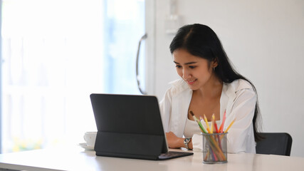 Smiling young asian woman designer concentrate working her project on computer tablet at creative workplace.