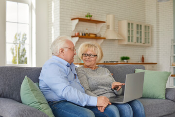 Smiling mature wife and husband looking at laptop screen sitting on sofa in cozy home. Senior couple relaxing at home watching videos or shopping online. Concept of the elderly and technology.