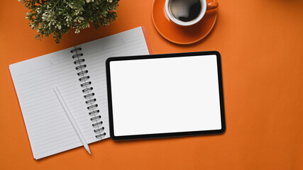 Mock up digital tablet with empty screen, notebook, coffee cup and plant on orange background.