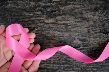 Top view of hand holding bright pink ribbon on dark wood background. Breast cancer awareness and...