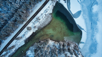 Birds eye view from a drone on a partial frozen lake in a winter landscape with trees and a road