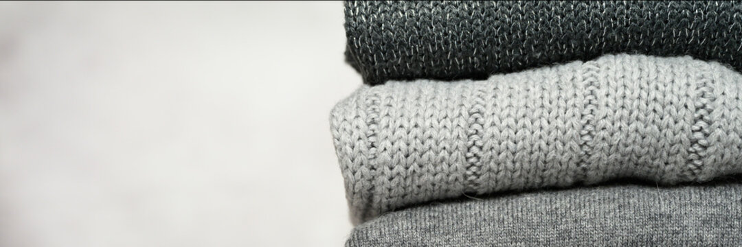 a stack of knitted winter sweaters in several shades of gray on gray background. banner