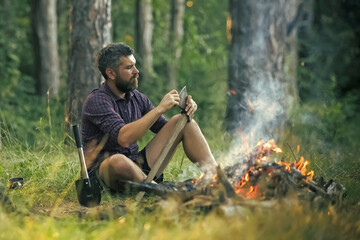 Man hipster sharpen axe. Camping, hiking, summer vacation. Guy relax at bonfire in forest. - obrazy, fototapety, plakaty