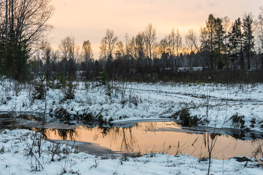 Winter landscape. The first snow fell, the sunset evening beautiful sky is reflected in the river. Blue evening shadows in the snow. Siberian nature.