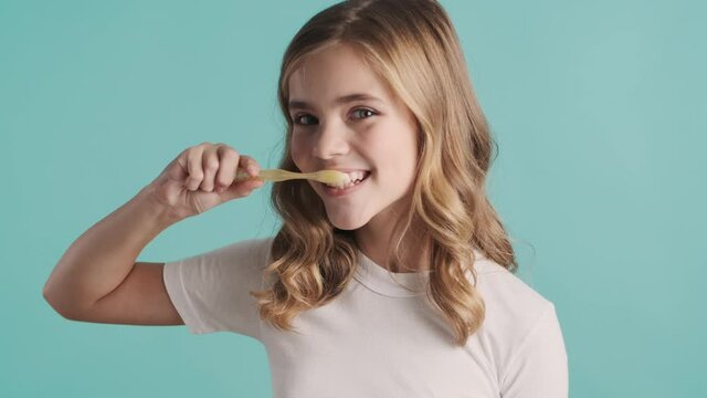 Portrait of beautiful blond teenager girl thoroughly brushing teeth recommending everyone to do this on camera over blue background