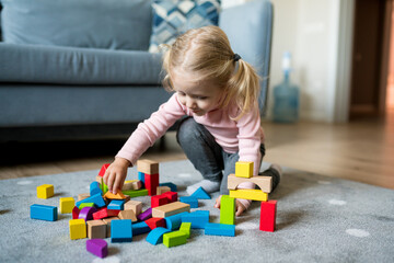 Cute Caucasian baby girl playing with colorful wooden blocks on the floor of the living room at home. Activities with children inside, early childhood development