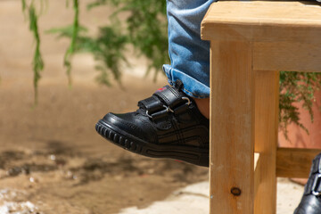 Baby feet of boy wears black leather shoes sitting on a wooden chair with copy space on the left 