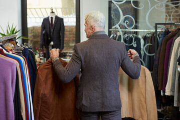 A photo from behind of a mature man with gray hair and a sporty physique is going holding suede jackets to the fitting room in a clothing store. A male customer with a beard wears a suit in a boutique