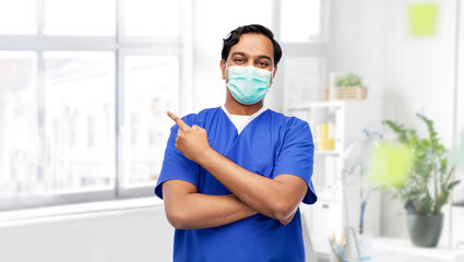 Fototapeta na wymiar healthcare, profession and medicine concept - indian doctor or male nurse in blue uniform and face protective medical mask showing something imaginary over medical office at hospital on background