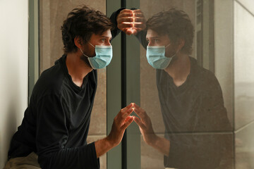 Sad man in isolation due to coronavirus world pandemic. Male in hotel quarantine wearing protective...