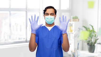 Fototapeta na wymiar healthcare, profession and medicine concept - indian doctor or male nurse in blue uniform, face mask for protection from virus disease and surgical gloves over medical office at hospital on background