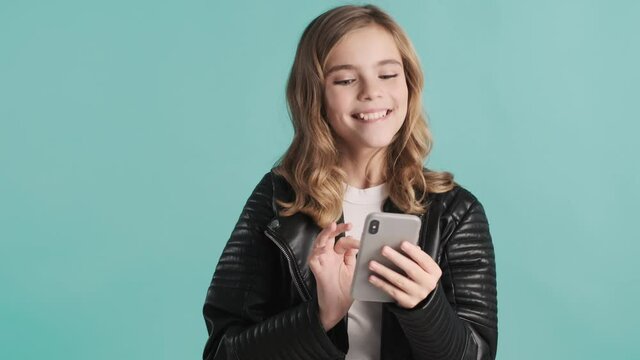 Cheerful blond teenager girl cheking social networks looking surprised over blue background. Modern technology concept