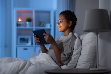 technology, internet and people concept - happy smiling young african american woman in glasses with tablet pc computer lying in bed at home at night