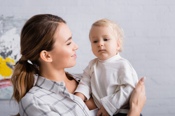happy young mother looking at little son while holding him at home