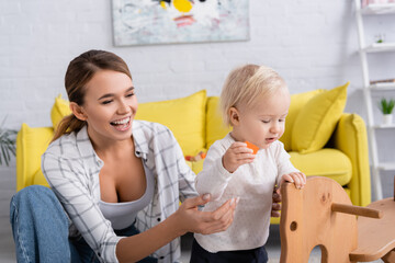 laughing woman supporting baby boy holding wooden cube near rocking horse