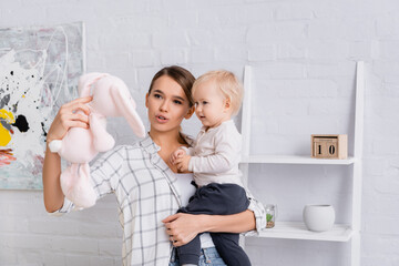 young mother holding baby boy and toy bunny at home