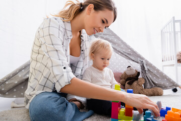 smiling woman playing with construction cubes with son on carpet near kids wigwam © LIGHTFIELD STUDIOS
